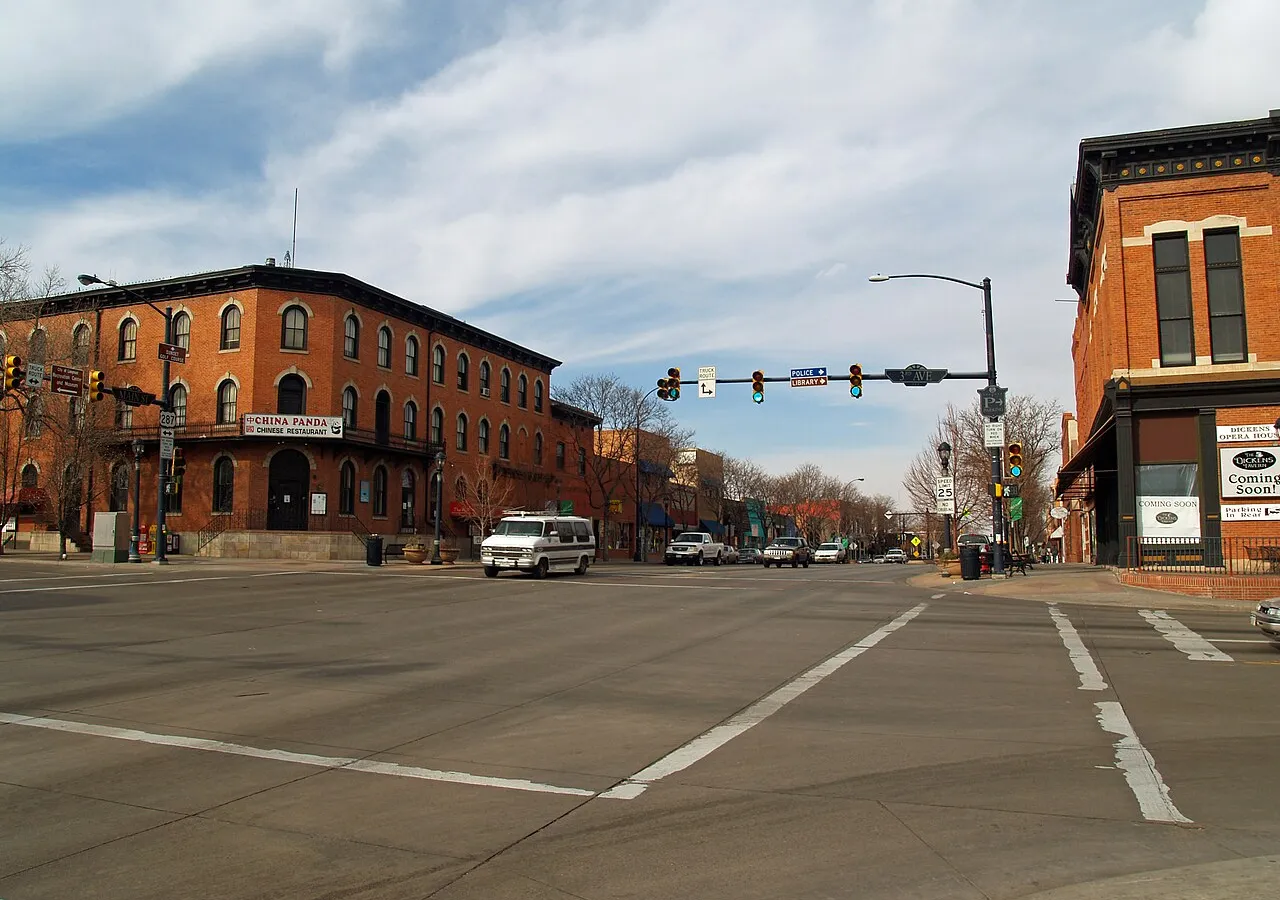 Main Street at 3rd Avenue in downtown Longmont on a late-winter day, the 1881 Dickens Opera House on the right corner and a three-storey red-brick block across the street, looking north up the 300 block under bare trees.
