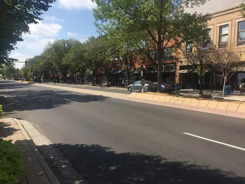 Main Street in downtown Longmont near 5th Avenue on a sunny September day, brick shopfronts under leafy street trees behind a planted median.
