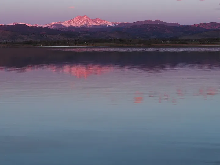 McIntosh Lake from its south shore at sunrise in March, the still water reflecting the pink first light on snow-covered Mount Meeker and the Front Range.