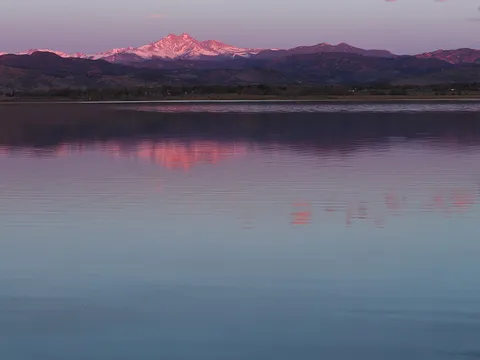 McIntosh Lake from its south shore at sunrise in March, the still water reflecting the pink first light on snow-covered Mount Meeker and the Front Range.