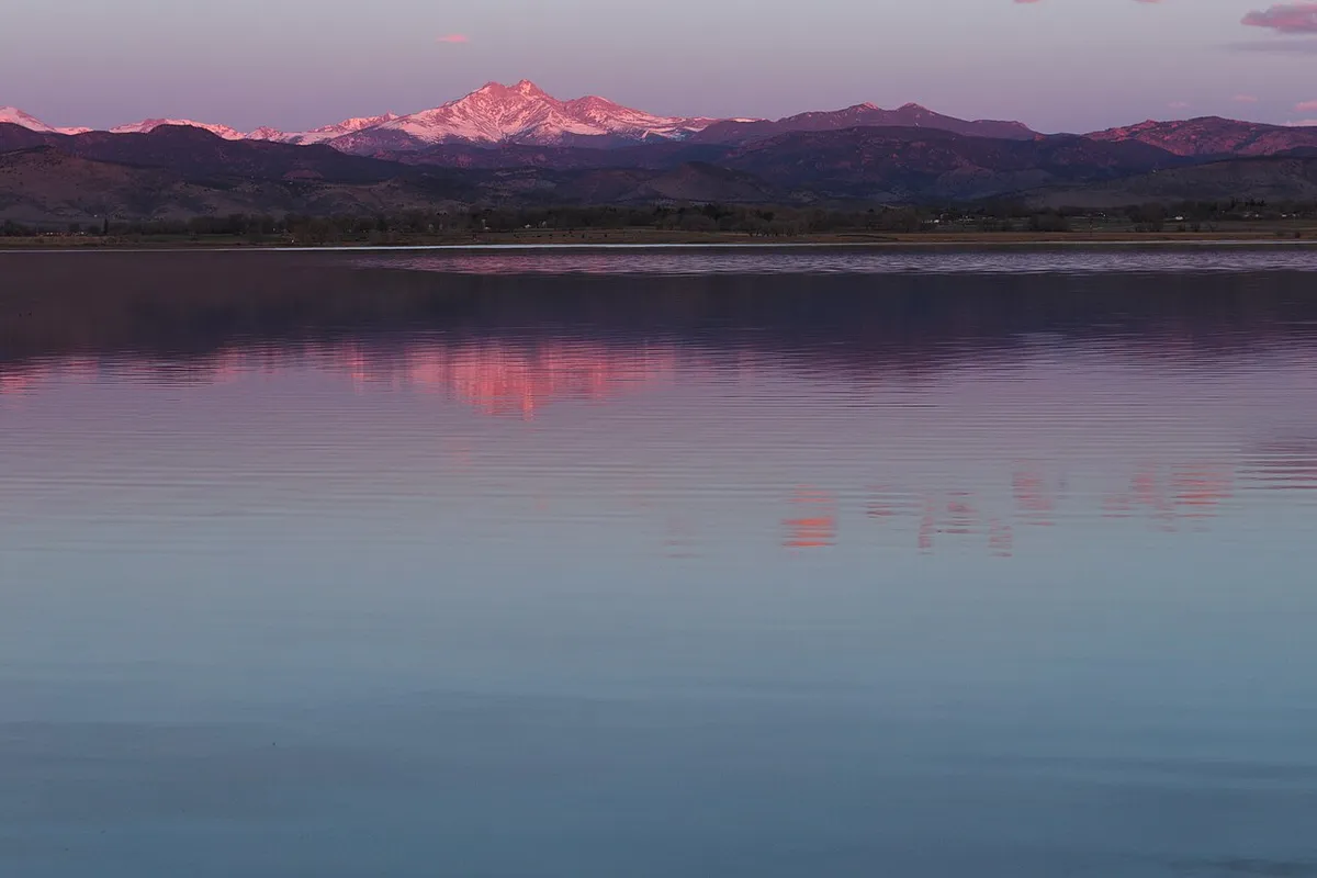McIntosh Lake from its south shore at sunrise in March, the still water reflecting the pink first light on snow-covered Mount Meeker and the Front Range.